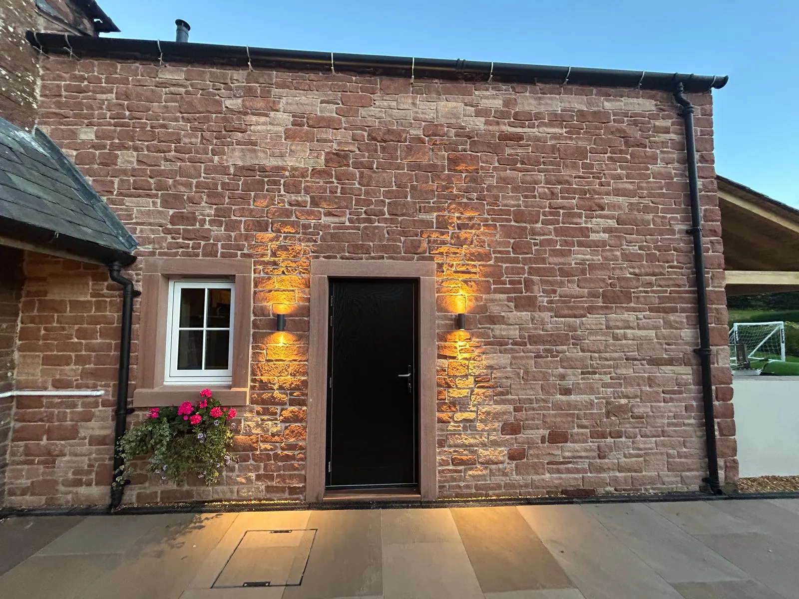 A house elevation in red sandstone at dusk, with a black door, two amber wall lights and a window box of pink geraniums